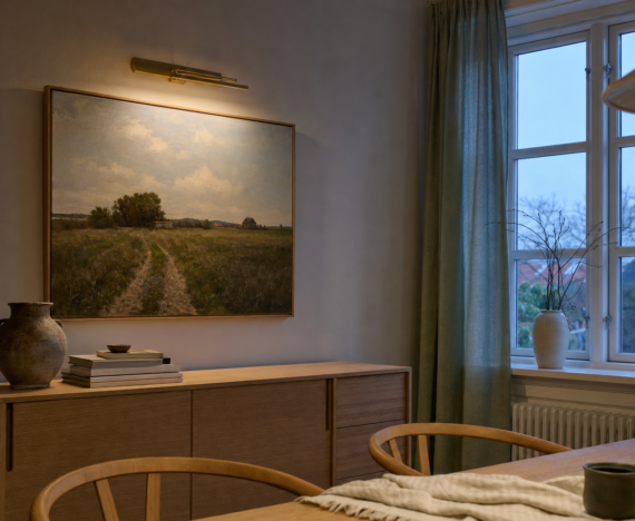 Landskabsmaleri over en egeskænk i en spisestue, oplyst af en smal messing-billedlampe i tusmørke med halvt trukne linnedgardiner ved vinduet