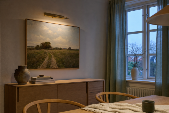 Landskabsmaleri over en egeskænk i en spisestue, oplyst af en smal messing-billedlampe i tusmørke med halvt trukne linnedgardiner ved vinduet